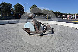 Old rusty anchor with chain placed in the middle of roundabout with traffic sign and concrete barrier.