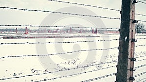 Old rusty barbed wire fence and distant ruined barracks of concentration camp in the snow. 4K steadicam shot