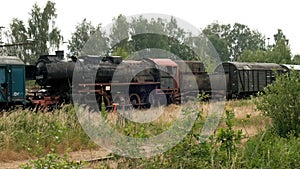 An old rusty black steam locomotive with a steam propulsion system and cars on abandoned tracks