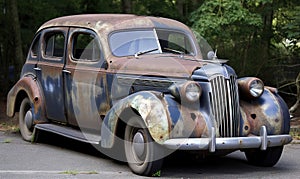 an old rusty car parked on the side of the road in a wooded area with trees in the backgrouds and a few bushes in the foreground