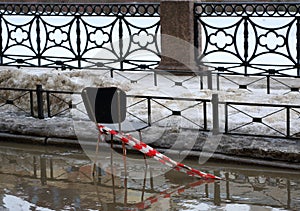 An old rusty chair with a piece of red and white ribbon stands in a puddle of meltwater on the embankment