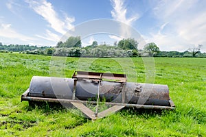 An old rusty farming roller lying in a field with a copse of bushes and trees in the far distance