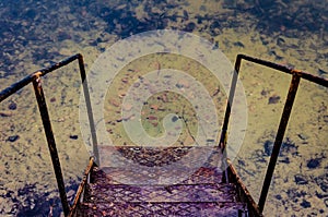 Old rusty iron stair near the water. Descent to the water. Pier near a lake or canal. View from above. Pure clear water and yellow