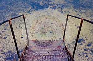 Old rusty iron stair near the water. Descent to the water. Pier near a lake or canal. View from above. Pure clear water and yellow