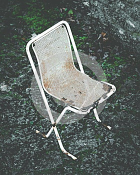 Old, rusty metal chair on stone background. Scenic, white, abandoned outdoor beach cafe furniture. Dark, moody, creepy, artistic