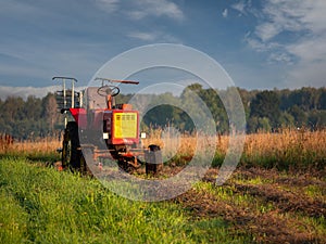 Old rusty tractor with grass cutting machine in a agriculture field with tall grass at stunning sunrise. Farming industry.