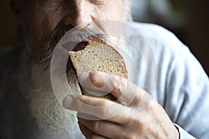 Old sad man with a long gray beard sitting by the table and eating bread