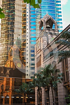 An old sandstone building with a bell tower is reflected in the windows of a glass skyscraper. Old colonial architecture and