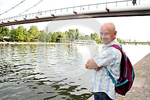 Old satisfied man, tourist with a backpack, stands at the bridge on the banks of the Main river in Frankfurt, looks at the