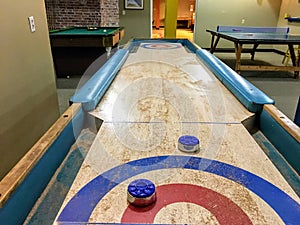 An old school shuffleboard table with blue shuffleboard pucks on top of it. The table is in a games room with a ping pong and pool