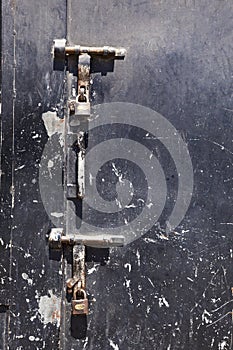 Old scratched black steel door with two large rusty latches and padlocks hanging from them.