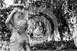 An old sculpture of a young woman carrying a water pitcher agains trees in monochrome colors