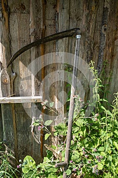 An old scythe stands against a wooden wall made of planks in the rays of sunlight