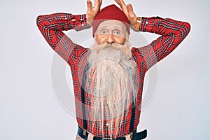 Old senior man with grey hair and long beard wearing hipster look with wool cap doing bunny ears gesture with hands palms looking