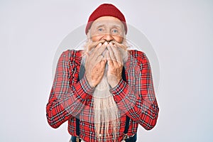 Old senior man with grey hair and long beard wearing hipster look with wool cap laughing and embarrassed giggle covering mouth