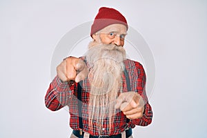 Old senior man with grey hair and long beard wearing hipster look with wool cap pointing fingers to camera with happy and funny