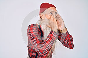 Old senior man with grey hair and long beard wearing hipster look with wool cap shouting angry out loud with hands over mouth