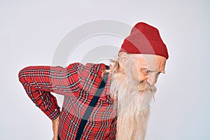 Old senior man with grey hair and long beard wearing hipster look with wool cap suffering of backache, touching back with hand,