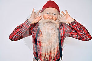 Old senior man with grey hair and long beard wearing hipster look with wool cap trying to hear both hands on ear gesture, curious
