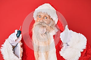 Old senior man with grey hair and long beard wearing santa claus costume holding car keys smiling happy and positive, thumb up