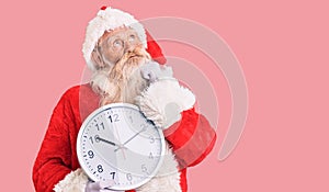 Old senior man with grey hair and long beard wearing santa claus costume holding clock serious face thinking about question with