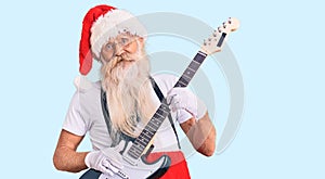 Old senior man with grey hair and long beard wearing santa claus costume playing electric guitar looking positive and happy