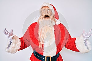 Old senior man with grey hair and long beard wearing santa claus costume with suspenders crazy and mad shouting and yelling with