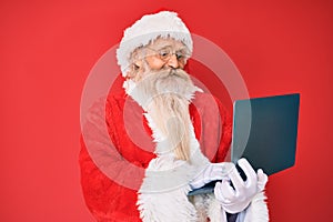 Old senior man with grey hair and long beard wearing santa claus costume using laptop smiling with a happy and cool smile on face