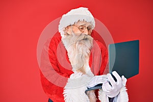 Old senior man with grey hair and long beard wearing santa claus costume using laptop smiling and laughing hard out loud because