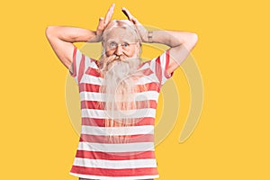 Old senior man with grey hair and long beard wearing striped tshirt doing bunny ears gesture with hands palms looking cynical and
