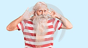 Old senior man with grey hair and long beard wearing striped tshirt trying to hear both hands on ear gesture, curious for gossip