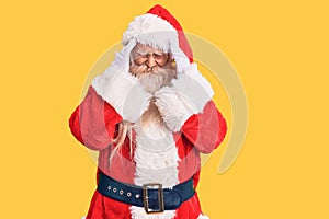 Old senior man with grey hair and long beard wearing traditional santa claus costume with hand on head, headache because stress