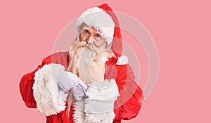 Old senior man with grey hair and long beard wearing traditional santa claus costume in hurry pointing to watch time, impatience,