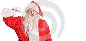 Old senior man with grey hair and long beard wearing traditional santa claus costume strong person showing arm muscle, confident