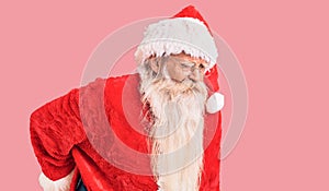 Old senior man with grey hair and long beard wearing traditional santa claus costume suffering of backache, touching back with