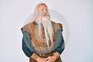 Old senior man with grey hair and long beard wearing viking traditional costume looking away to side with smile on face, natural