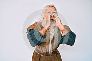 Old senior man with grey hair and long beard wearing viking traditional costume shouting angry out loud with hands over mouth