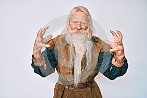 Old senior man with grey hair and long beard wearing viking traditional costume shouting frustrated with rage, hands trying to