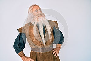 Old senior man with grey hair and long beard wearing viking traditional costume smiling looking to the side and staring away