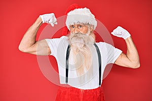 Old senior man with grey hair and long beard wearing white t-shirt and santa claus costume showing arms muscles smiling proud