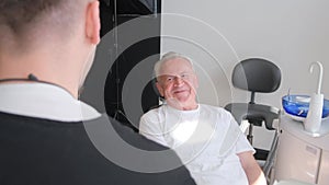 An old senior man sitting in a dental chair in a dentist's office, he is talking to a doctor.