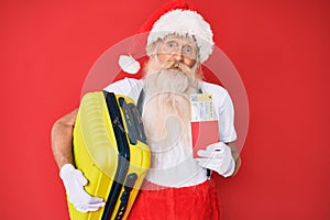Old senior man wearing santa claus costume holding suicase and boarding pass clueless and confused expression