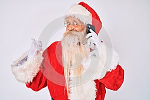 Old senior man wearing santa claus costume speaking on the phone pointing thumb up to the side smiling happy with open mouth