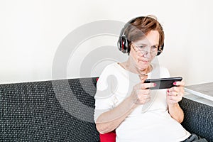 Old senior woman female sit home flat couch hold smartphone play game happy headphones alone relax enjoy