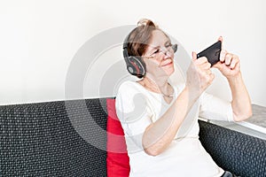 Old senior woman female sit home flat couch hold smartphone play game happy headphones alone relax enjoy