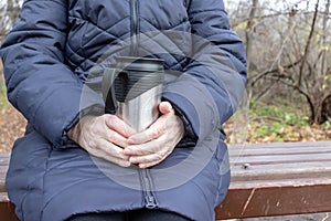 An old senior woman sitting on a bench in late autumn park and holding thermo cup in hands, hot drinks outdoor concept