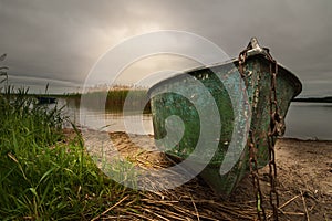 old shabby fishing boat on a chain on the lake with a reed against a dramatic cloudy sky with a glow. picturesque summer landscape