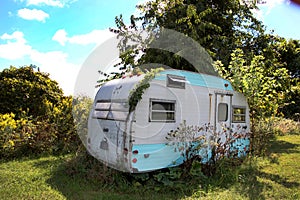 Old small camper trailer with overgrown weeds stuck in the ground.