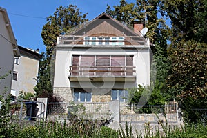 Old small family house with large upper balcony and dilapidated closed wooden window blinds surrounded with stone and metal fence