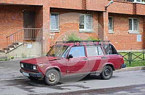 Old Soviet rusty burgundy car stands near a red brick house
